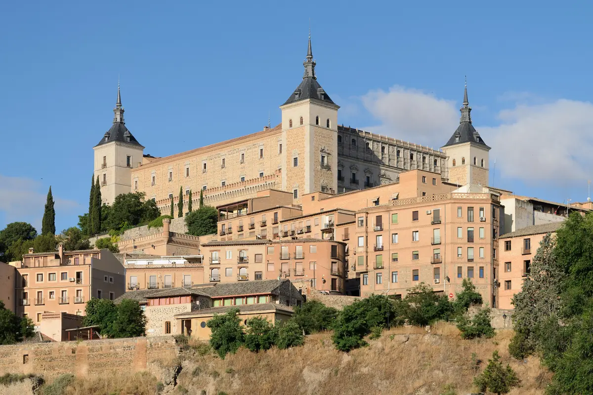 The Alcázar of Toledo, a large stone building with corner towers topped by dark spires, above terracotta-roofed houses on the hillside