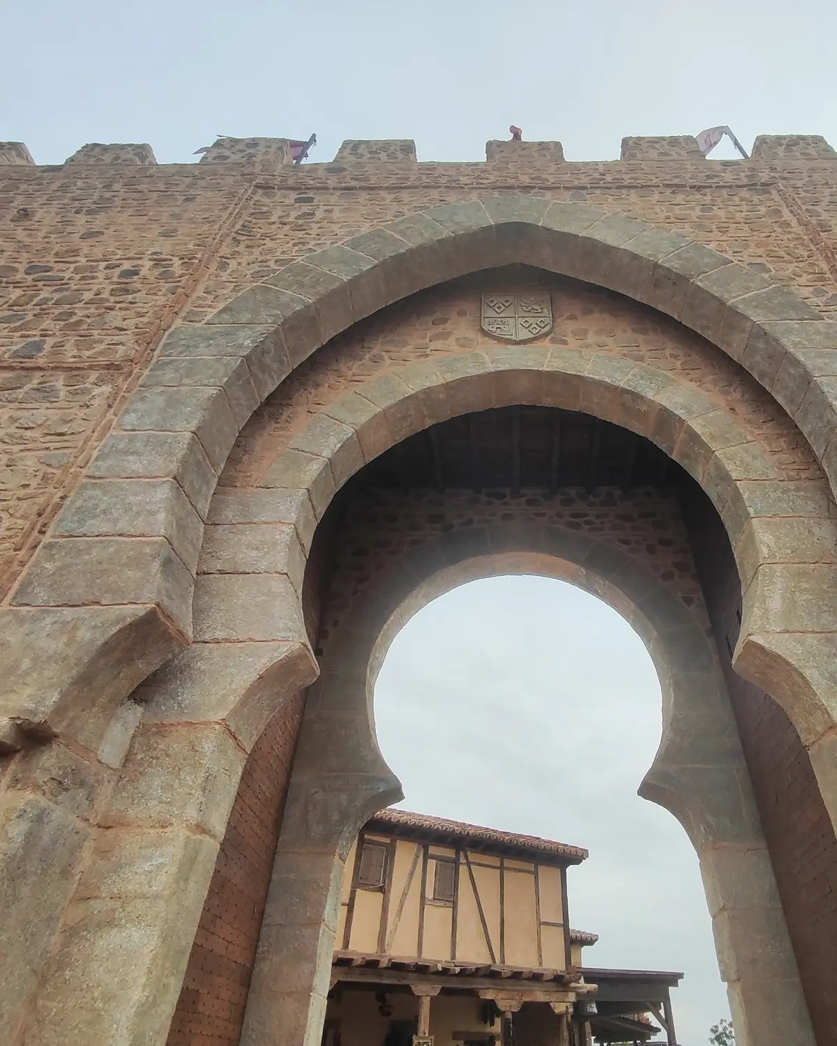 A tall stone gateway with a keyhole-shaped horseshoe arch and battlements, with a half-timbered house seen through the opening