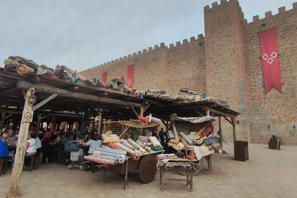 Market stalls piled with cloth and goods under a timber roof, beside a high crenellated wall hung with a red banner, with visitors seated in the shade