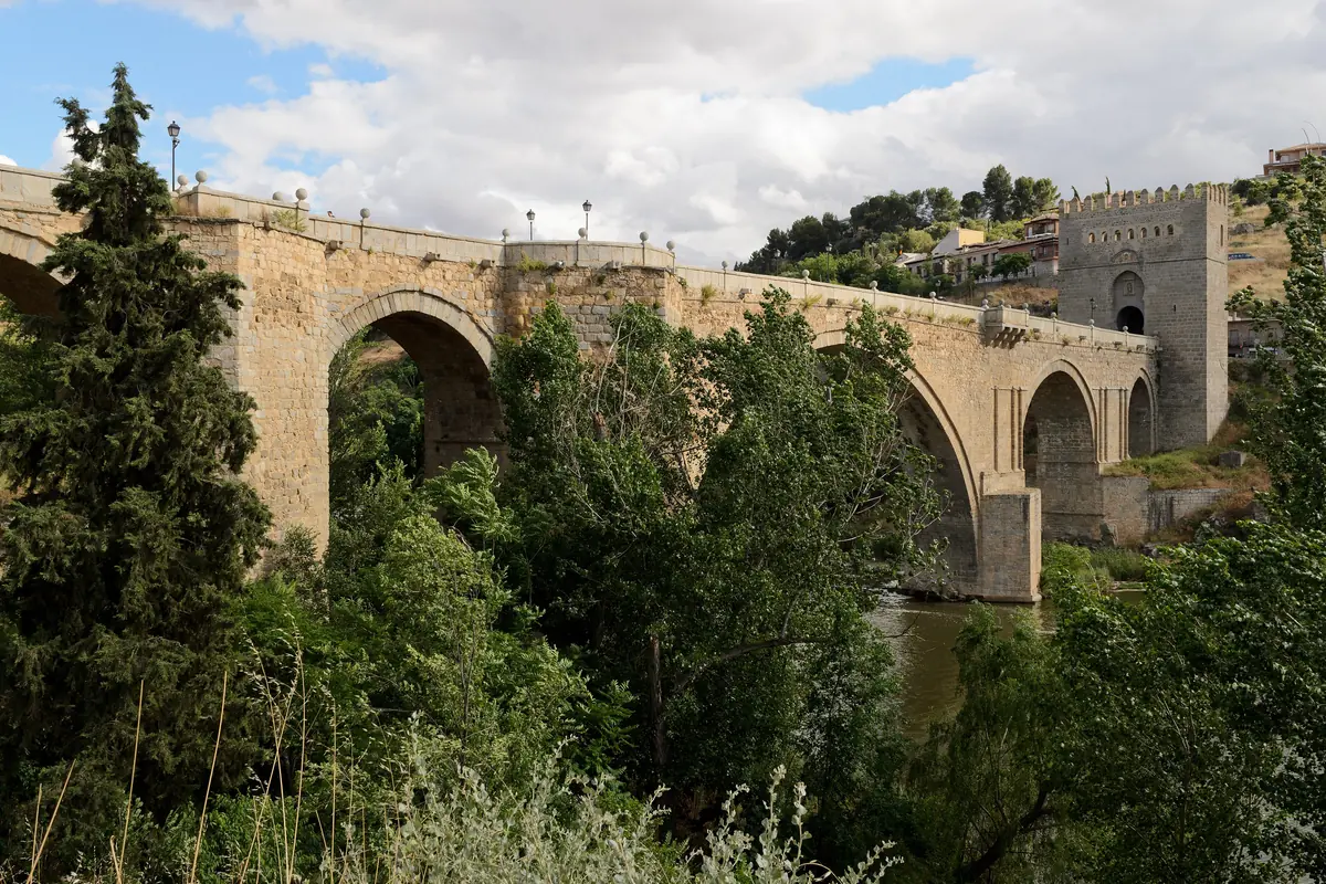 The stone San Martín Bridge seen through trees, with arches over the Tagus and a fortified tower at the far end