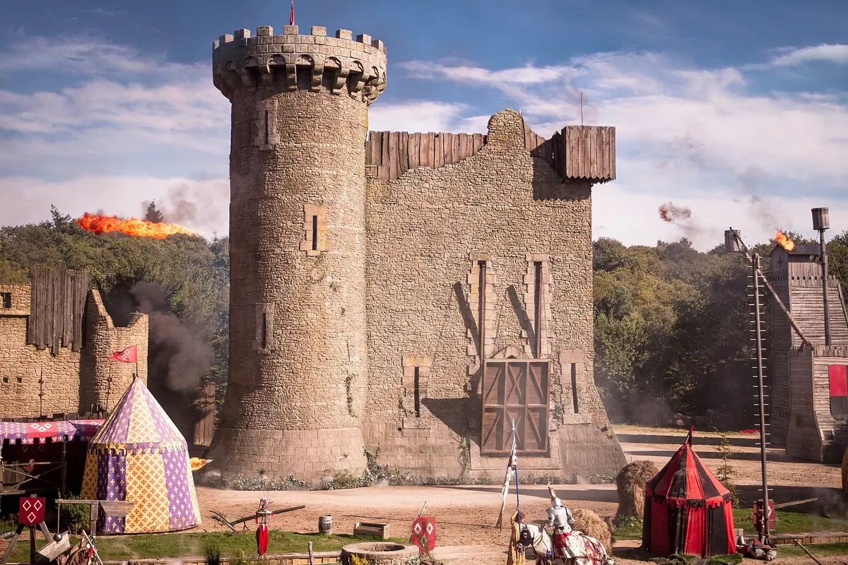 A stone round tower and ruined castle wall in an arena, with striped medieval tents, a mounted knight in white and flames and smoke from a catapult in the background