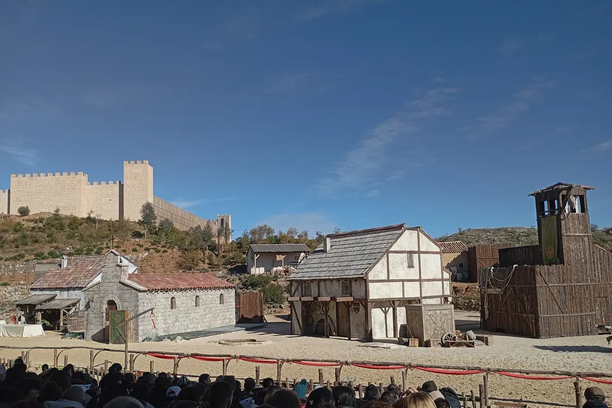 Spectators in the stands face a village set of half-timbered houses and a wooden tower, with a stone castle wall on the hill behind