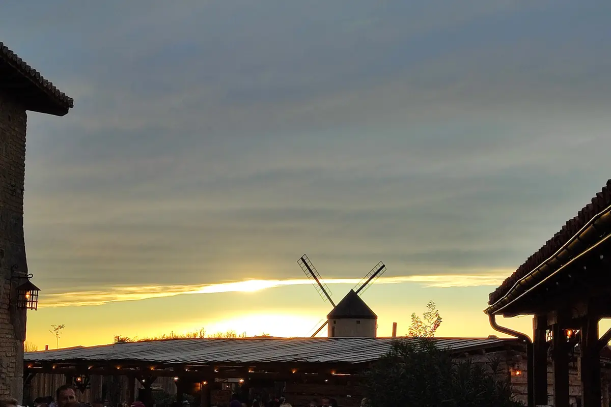 A windmill silhouetted on the skyline at dusk, with tiled roofs on either side and a band of orange light under grey cloud