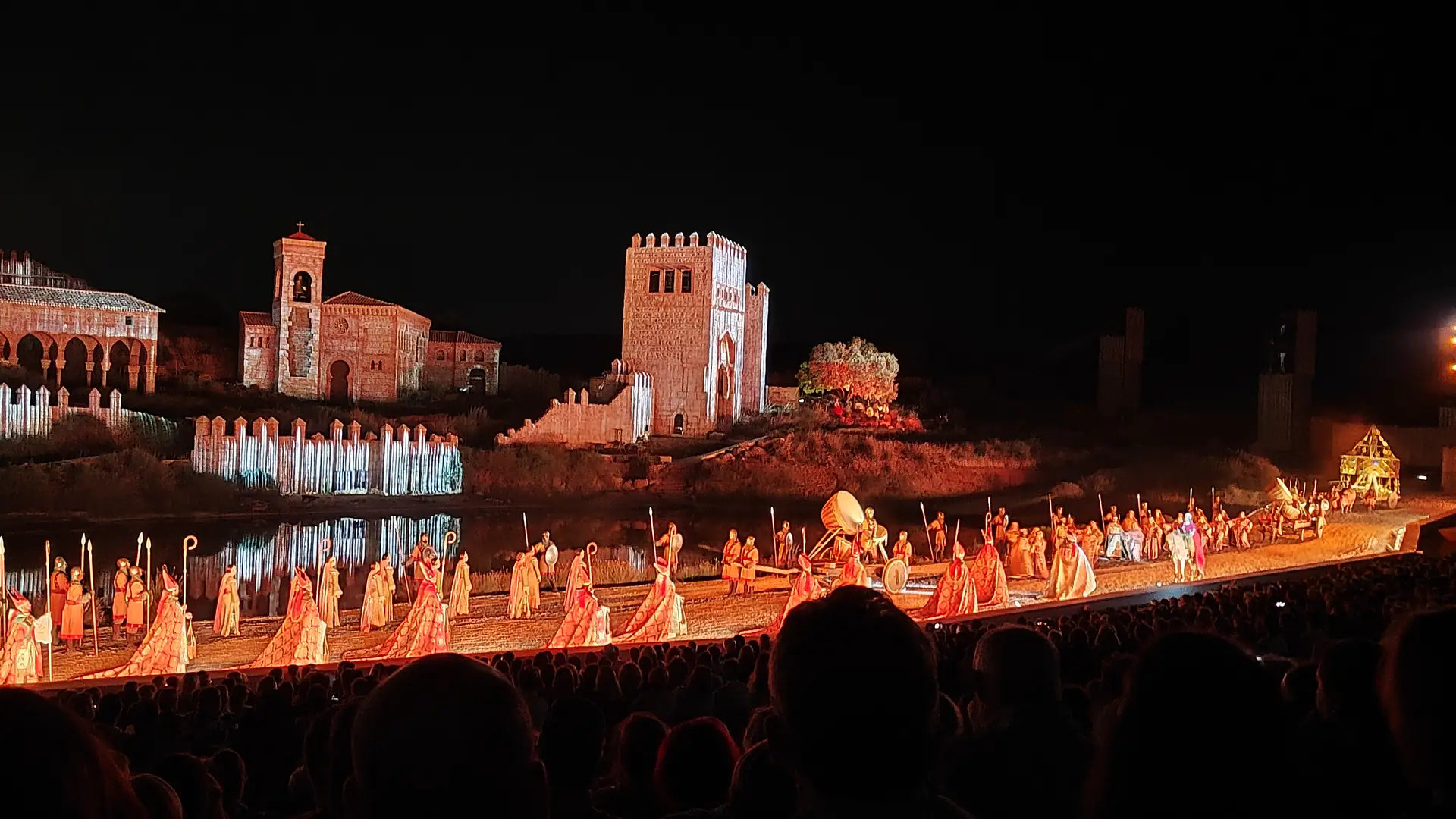 The El Sueño de Toledo night show: a long line of performers in orange and gold costumes walks along the stage edge before floodlit stone towers and a church that reflect in the water, with the audience's heads in the foreground.
